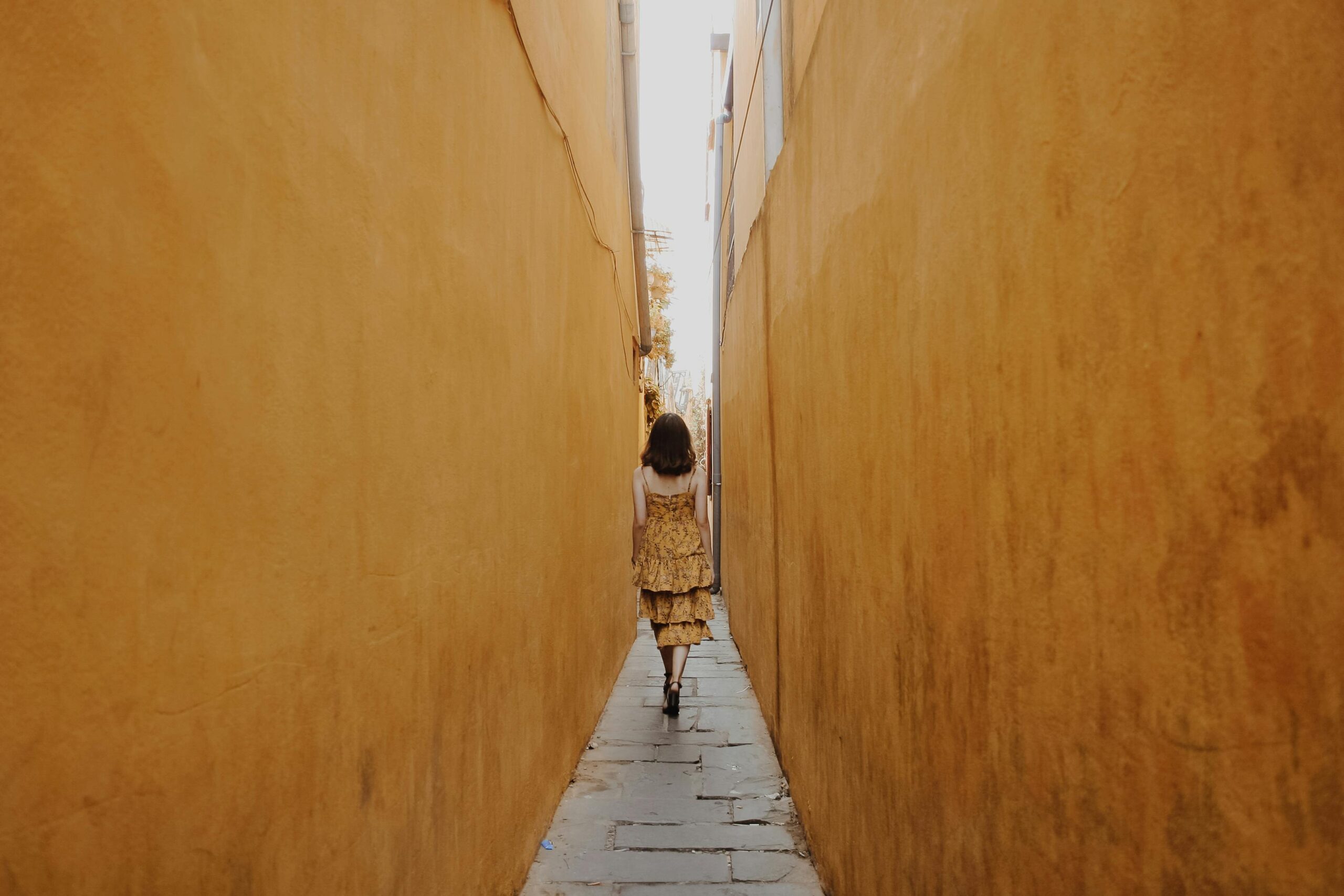 Woman walking down an alleyway with high yellow walls on either side, representing trauma keeping you stuck.